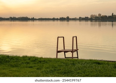 A serene lakeside view with an empty wooden chair at sunset, evoking solitude and reflection during summer - Powered by Shutterstock