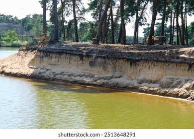 A serene lakeside view featuring a sandy bank with exposed layers of soil, surrounded by tall trees. The calm water reflects the greenery, creating a peaceful natural scene. - Powered by Shutterstock