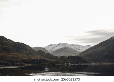 A serene landscape of a boat on a calm lake surrounded by snow-capped mountains under an overcast sky. - Powered by Shutterstock