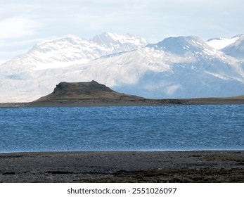 A serene landscape featuring a calm lake with a small island in the foreground, surrounded by majestic snow-capped mountains in the background under a cloudy sky. - Powered by Shutterstock