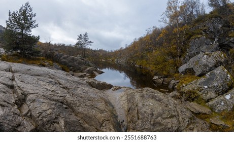 A serene landscape featuring a small, calm lake surrounded by rocky terrain and sparse trees with autumn foliage under an overcast sky. - Powered by Shutterstock