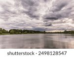 A serene landscape with a lake, trees, and cloudy skies. The scene exudes tranquility as the still water reflects the overcast sky. Rustling leaves and a mix of tree types add to nature s calm beauty.