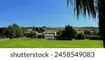 A serene landscape, with a lush green field and a small cricket pavilion, in the background are  rolling hills and a clear blue sky in, Bradley, Keighley, UK