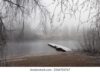 A serene landscape showcases a foggy morning by the lake, featuring a wooden dock extending into the water and surrounding trees cloaked in mist. Distant houses fade into the background. - Powered by Shutterstock