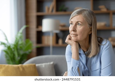 A serene mature woman in a blue shirt stares thoughtfully while sitting in a well-lit living room. The peaceful atmosphere highlights her pensive mood. - Powered by Shutterstock