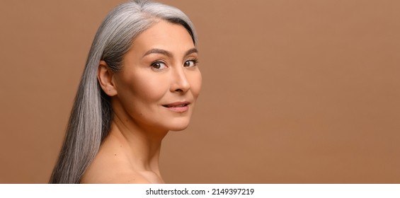 Serene middle-aged Asian woman with fresh clean healthy skin isolated on grey. Well-looking mature korean lady looks at camera, small wrinkles near eyes. Anti aging care, natural beauty