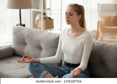 Serene Millennial Woman Sitting On Couch In Living Room At Home. Girl Take A Break For Meditation Yoga Practice. Thinking, Stress Relief, Healthy Good Habits, Mental Health, Mindful Lifestyle Concepts