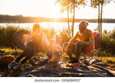 A serene moment captured as a bearded senior man, a young woman, and a child unwind on camping chairs by a lake, surrounded by untouched nature, basking in the golden sunset glow. Family Enjoying - Powered by Shutterstock