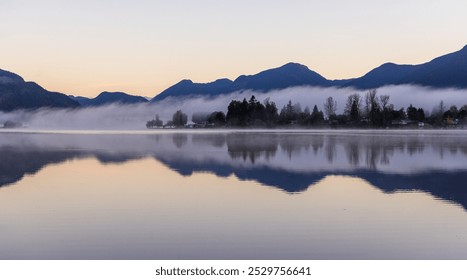 A serene morning view with mist floating over a calm lake in Mission, BC, Canada, reflecting the surrounding mountains. Peaceful and tranquil atmosphere captured at dawn. - Powered by Shutterstock