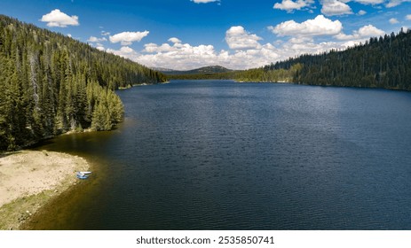 A serene mountain lake with calm water and lush pine forests under a vibrant blue sky with fluffy clouds, offering a peaceful natural retreat. - Powered by Shutterstock