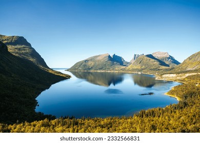 Serene mountain lake nestled amidst lush green trees and surrounded by majestic mountains.Crystal-clear blue water reflects clear sky with few fluffy clouds
