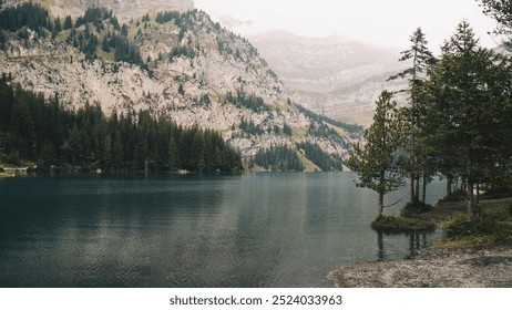 A serene mountain in Switzerland, Kanderstag, with the lake Oeschinensee, surrounded by lush pine trees and rocky cliffs under a overcast sky. - Powered by Shutterstock