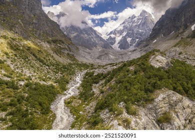 A serene mountain valley with a flowing stream, surrounded by rugged terrain and lush greenery. Snow-capped peaks rise in the background under a partly cloudy sky. - Powered by Shutterstock