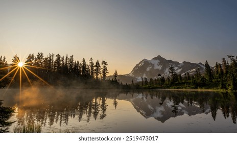 A serene mountainous landscape at sunrise with a sunburst through tall evergreen trees reflecting on a calm, misty lake. Snow-capped peaks and a clear blue sky form the backdrop. - Powered by Shutterstock