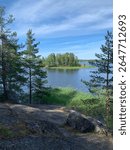 A serene natural landscape featuring a calm lake surrounded by lush green pine trees and rocky foreground. Mon Repos Park. Vyborg. Russia.