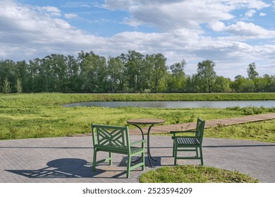 Serene outdoor scene. Two green wooden chairs and round table set. alm pond , surrounded by lush greenery. Wooden pathway beside water, tranquil atmosphere. Shishkin Ponds Park, Yelabuga, Russia - Powered by Shutterstock