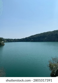 A serene panoramic view of a tranquil, deep blue-green lake reflecting the vast expanse of the sky, bordered by dense, verdant forests and rocky outcrops