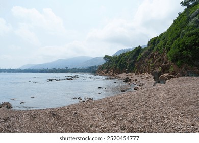 A serene pebble beach bordered by a lush hillside, with distant mountains and calm sea under a partly cloudy sky. A peaceful coastal landscape inviting relaxation and exploration - Powered by Shutterstock