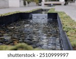 serene photo of a small man-made pond with a gentle mini waterfall cascading into it. The water flows over smooth stones, surrounded by a strip of moss. This creates a peaceful zen atmosphere.