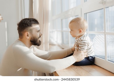 Serene portrait of father baby interaction by window, with dad attentive gaze and little son curious exploration emphasizing familial connection. - Powered by Shutterstock