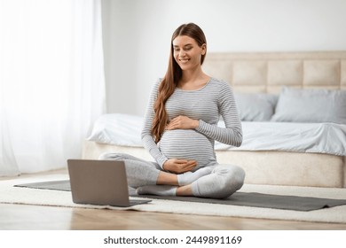 A serene pregnant woman sits cross-legged on a yoga mat in her bedroom, in front of a laptop, practicing prenatal yoga - Powered by Shutterstock