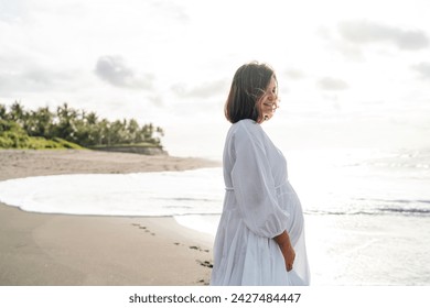 A serene profile of pregnant woman on beach, basking in gentle glow of sun with soft smile, her white dress billowing slightly in the sea breeze, epitomizing peace and maternal anticipation - Powered by Shutterstock