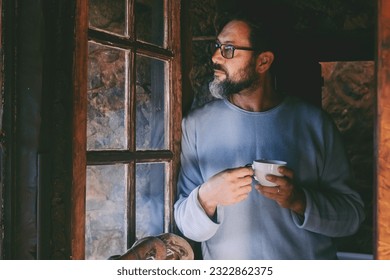 Serene and relaxed mature man standing indoor at home near the window and looking outside thinking. Real people lifestyle. Male with beard and glasses drinking coffee. Wooden chalet vacation winter - Powered by Shutterstock