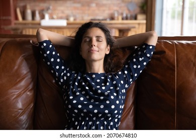 Serene rest. Peaceful young latina female relax on comfy couch alone with hands over head keep eyes closed. Happy tranquil millennial lady enjoy breathing fresh conditioned air at home feel no stress