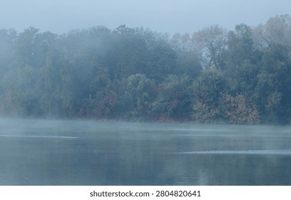 A serene river flows gently through a landscape of trees on a misty autumn morning, creating a tranquil and atmospheric view