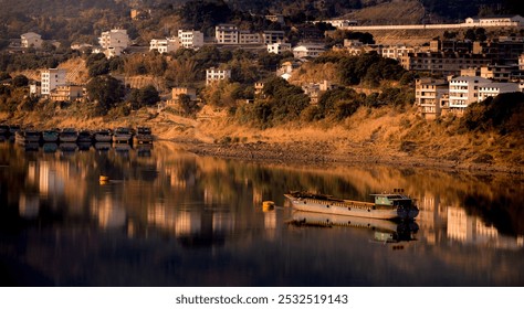 A serene river scene with a solitary barge floats gently. Hillside buildings and greenery reflect on the calm water at sunset, creating a tranquil ambiance. - Powered by Shutterstock