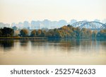 A serene riverscape with autumnal trees reflected in the water, a bridge in the distance, and a hazy city skyline. The calm atmosphere evokes the transition between nature and urban life.