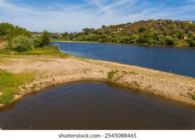 A serene riverside landscape with gently sloping sandy banks, lush greenery, and a calm river flowing alongside a hilly, tree-covered countryside under a clear blue sky - Powered by Shutterstock