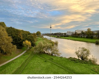 A serene riverside landscape at sunset, with lush greenery, a winding pathway, and a cityscape in the background under a colorful, cloud-streaked sky. - Powered by Shutterstock