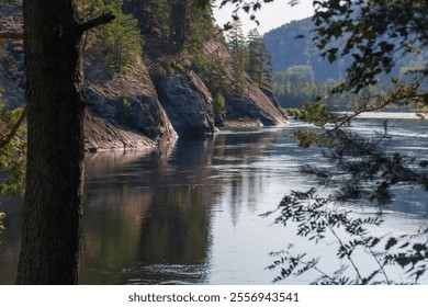 A serene riverside scene framed by trees, featuring calm waters reflecting rocky cliffs and lush green forests in soft sunlight. - Powered by Shutterstock