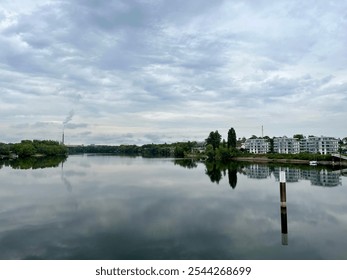 A serene riverside scene with modern apartment buildings and a distant industrial chimney emitting smoke, reflected in the calm water under an overcast sky. - Powered by Shutterstock