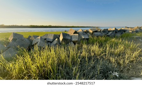 A serene riverside scene at sunrise with traditional boats docked on the shore, surrounded by lush green grass and concrete blocks. - Powered by Shutterstock