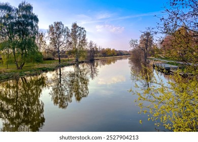 A serene riverside view with trees lining the riverbanks, reflecting perfectly in the calm water under a colorful sunset sky. The image captures the tranquility of nature and the peaceful atmosphere. - Powered by Shutterstock