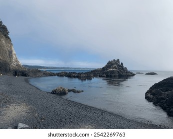 A serene, rocky beach with dark pebbles and rugged cliffs overlooks calm ocean waters under a cloudy sky. - Powered by Shutterstock