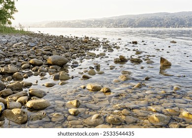 A serene rocky shoreline with calm waters and a distant view of hills under a clear sky. - Powered by Shutterstock