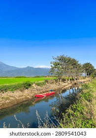 Serene rural landscape of Central Java with a traditional red boat on a calm irrigation canal, lush green rice fields, and a majestic mountain range under a clear blue sky, Indonesia.