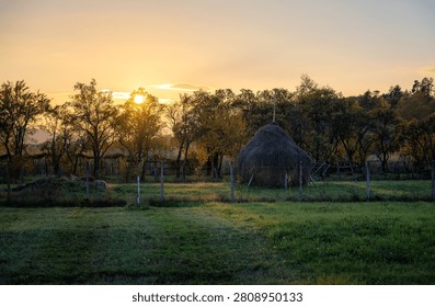 Serene rural landscape with a haystack and trees at sunset, casting a warm glow over the countryside.