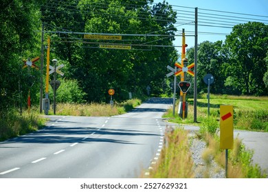 A serene rural road features bright railway crossing signals amidst vibrant greenery under clear blue skies. The area is peaceful, inviting exploration - Powered by Shutterstock