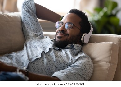 Serene Satisfied Young Adult African American Man Lying On Comfortable Sofa Wear Wireless Headphones Enjoy Listen Modern Music Audio Book With Eyes Closed Meditate Relax Feel No Stress Chill At Home