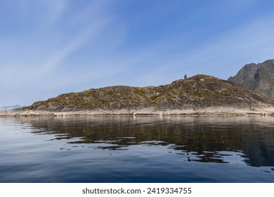 A serene Scandinavian fjord with a lone lighthouse atop a moss-covered rocky island, its reflection on the calm sea water under a clear blue sky. Lofoten, Norway - Powered by Shutterstock
