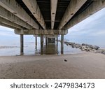 A serene scene beneath a pier on a beach during low tide, featuring rocks and a cloudy sky overhead