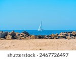 Serene scene at Venice Beach featuring a sandy shoreline, scattered boulders, a tranquil ocean, and a sailboat in the distance.