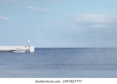 Serene seascape with a calm blue ocean under a clear sky, featuring a white pier with green lighthouse, red buoy, distant sailboat, and buoy line. Ideal for peaceful maritime themes and nautical back - Powered by Shutterstock