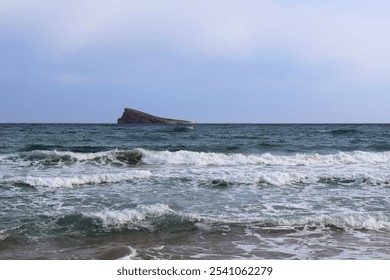 Serene seascape with gentle waves lapping onto a sandy beach, a rocky island visible in the calm blue-green ocean. Overcast sky adds a tranquil, contemplative atmosphere to the coastal scene. - Powered by Shutterstock