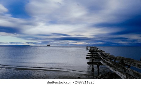 A serene seascape at Punta Arenas, Chile, featuring an old wooden pier extending into the calm waters with a ship on the horizon under a dramatic, cloudy sky.
 - Powered by Shutterstock