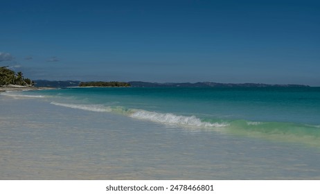 A serene seascape. The waves of the aquamarine ocean foam and spread over the sandy beach. A small island  with tropical vegetation, in the distance. Mountains against a clear blue sky. Copy space.  - Powered by Shutterstock